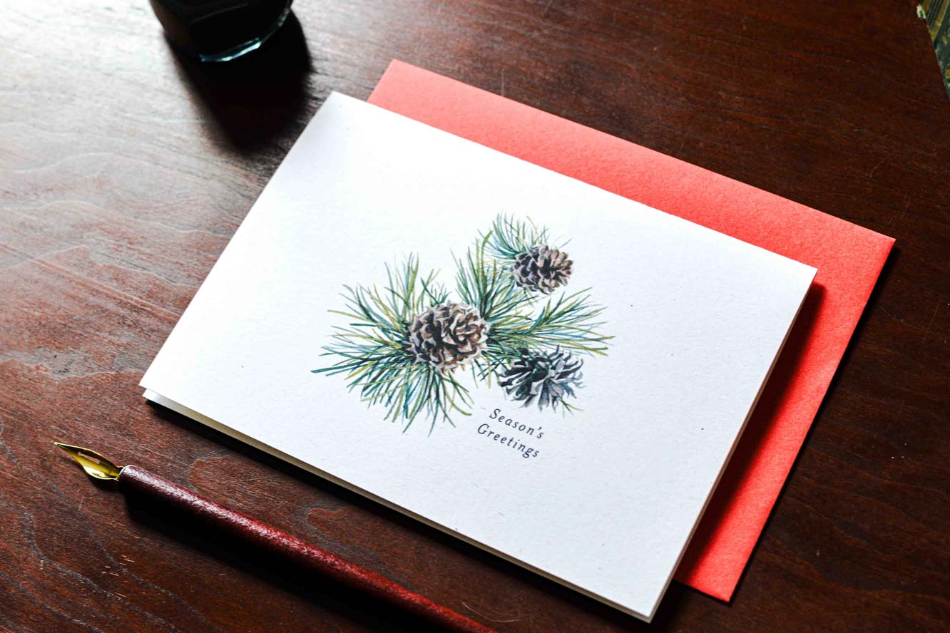 A white greeting card with a watercolor illustration of pine needles and pinecones, labeled 'Season's Greetings,' placed on top of a coral-red envelope on a dark wooden table. A fountain pen is visible at the lower left.