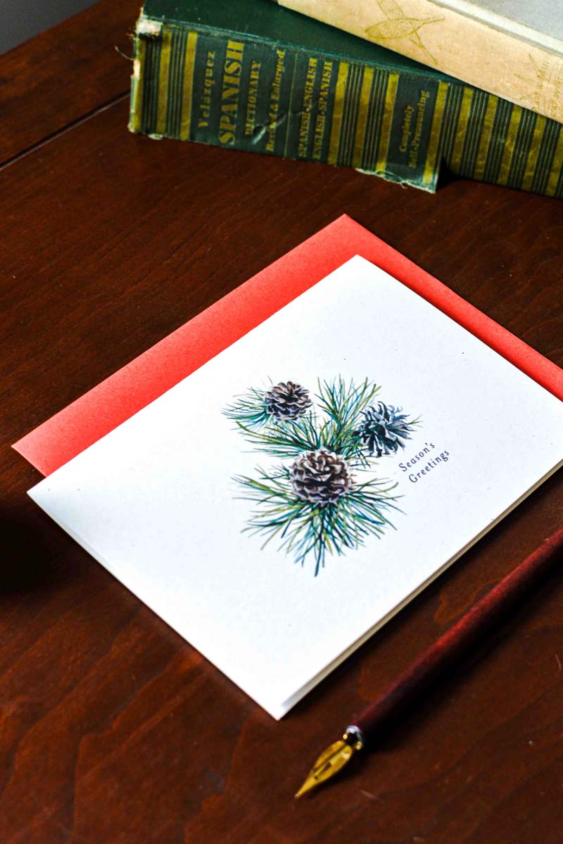 A square white greeting card with a watercolor pine branch and pinecone design, placed on a red envelope on a dark wooden table. Vintage books and a fountain pen are visible in the background.