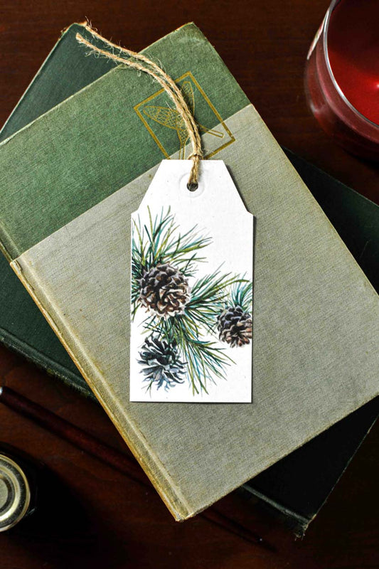 A white paper gift tag with a pine branch and pinecone illustration, attached with twine to an old green book on a dark wooden surface.
