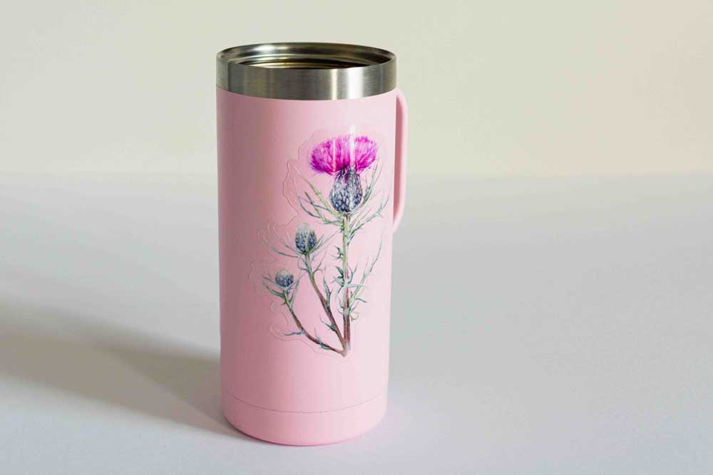 Pink travel mug with clear thistle sticker on a light gray background.