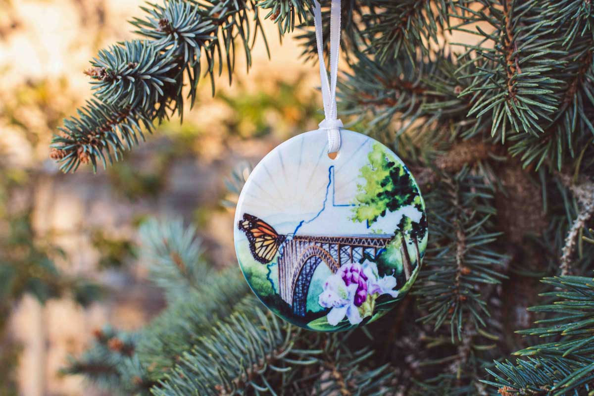 Decorative WV ornament with the New River Gorge Bridge, state outline, butterfly and tree design hanging on a pine branch.
