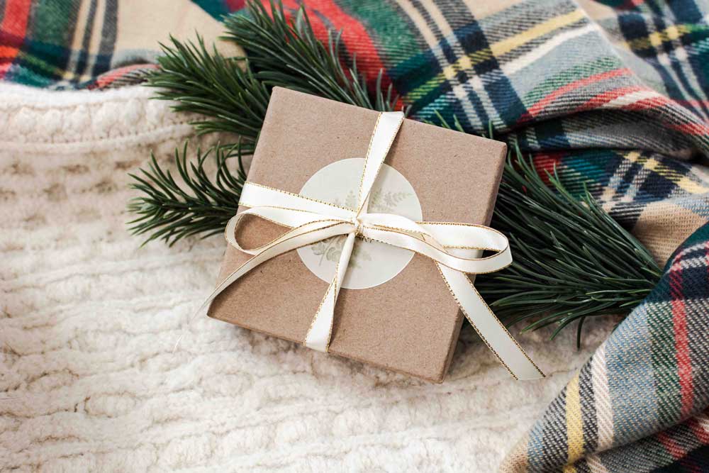 Gift box with a white ribbon on a textured blanket with plaid fabric and greenery.