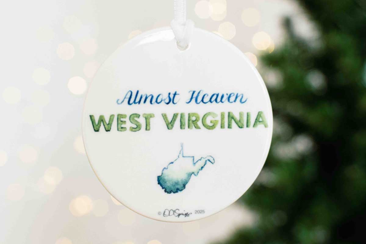 Round ornament with 'Almost Heaven West Virginia' text and outline of West Virginia on a blurred Christmas tree background.