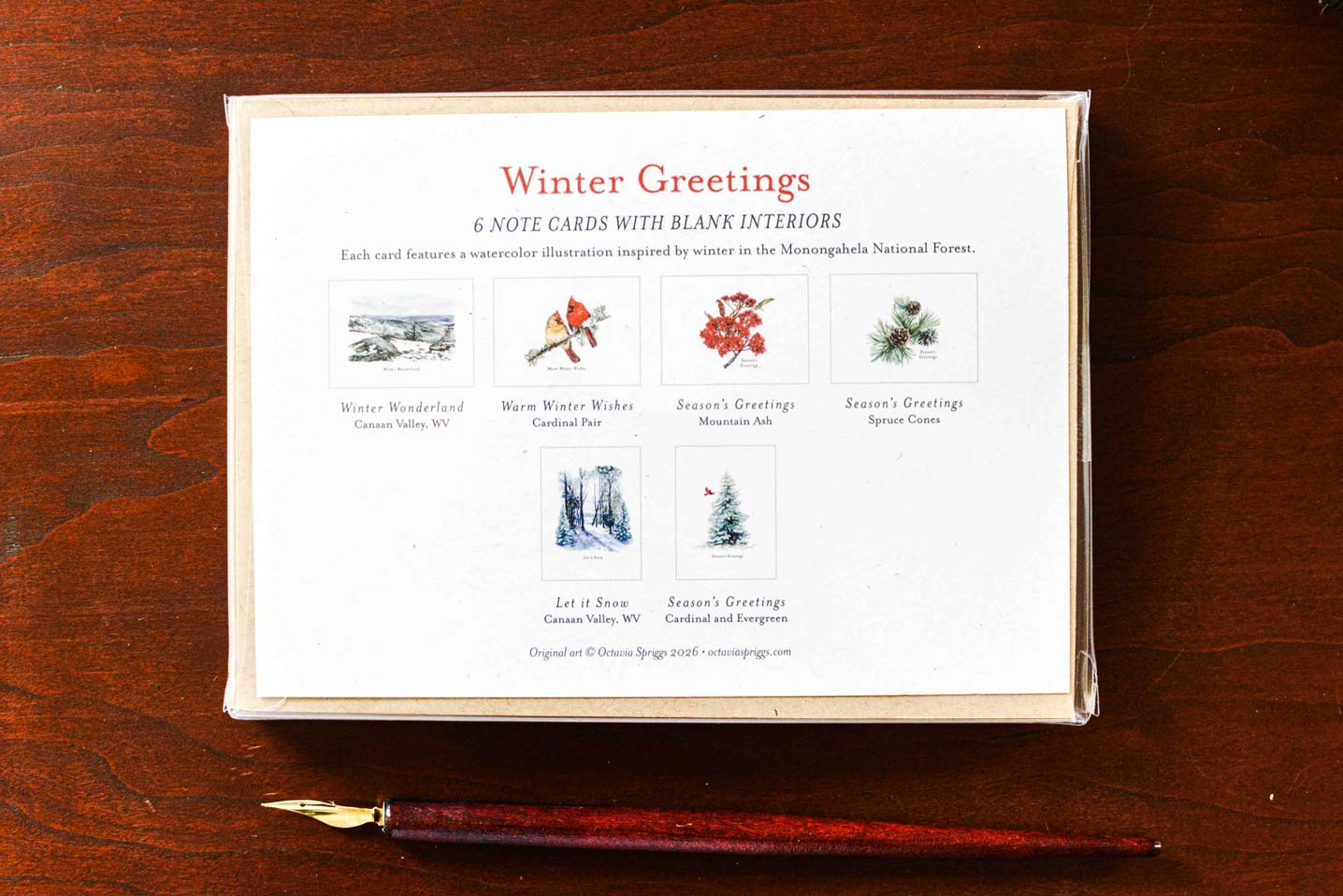 A boxed set of winter-themed note cards placed on a dark wooden surface, with a fountain pen lying in front. The front of the box reads 'Winter Greetings' and shows small illustrated cards with seasonal scenes and text.
