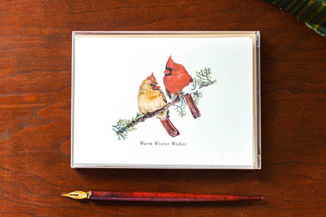 A greeting cards with a white background featuring an illustration of two cardinals on a branch, with the text 'Warm Winter Wishes' below. The card is shown in a clear box on a dark wood surface next to a gold-tipped pen.
