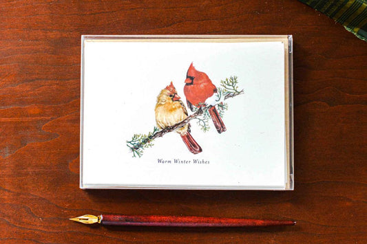 A greeting cards with a white background featuring an illustration of two cardinals on a branch, with the text 'Warm Winter Wishes' below. The card is shown in a clear box on a dark wood surface next to a gold-tipped pen.
