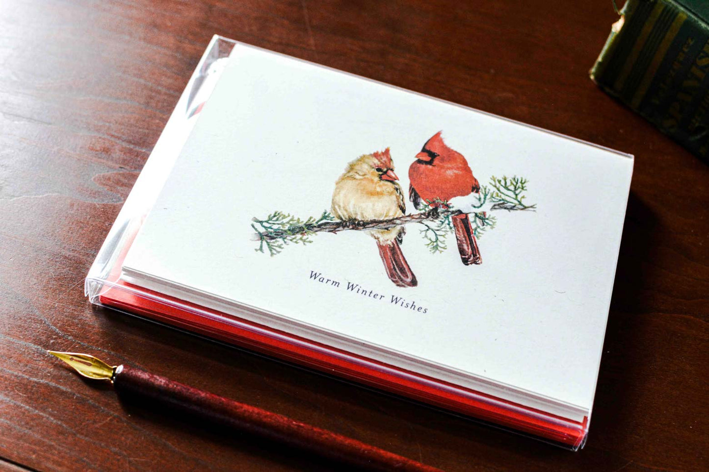A stack of white greeting cards with a watercolor illustration of two cardinals on a branch and the text 'Warm Winter Wishes,' photographed on a wooden table next to a fountain pen.