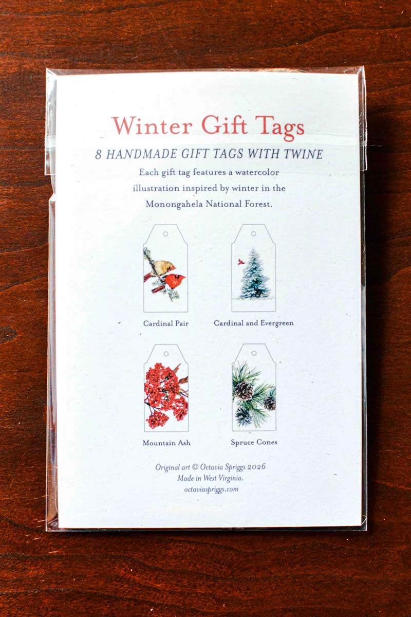 A clear plastic sleeve containing a set of winter-themed gift tags on a dark wooden tabletop. The back of the tags shows printed illustrations and names including Cardinal Pair, Cardinal and Evergreen, Mountain Ash, and Spruce Cone.