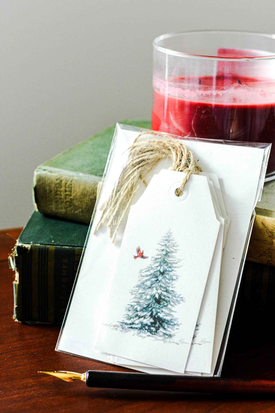 A clear plastic sleeve holds a white gift tag printed with a snowy evergreen tree and a small red bird, tied with twine. The tag is styled on a wooden surface with stacked vintage green books, a glass of red beverage, and a black pen in the background.