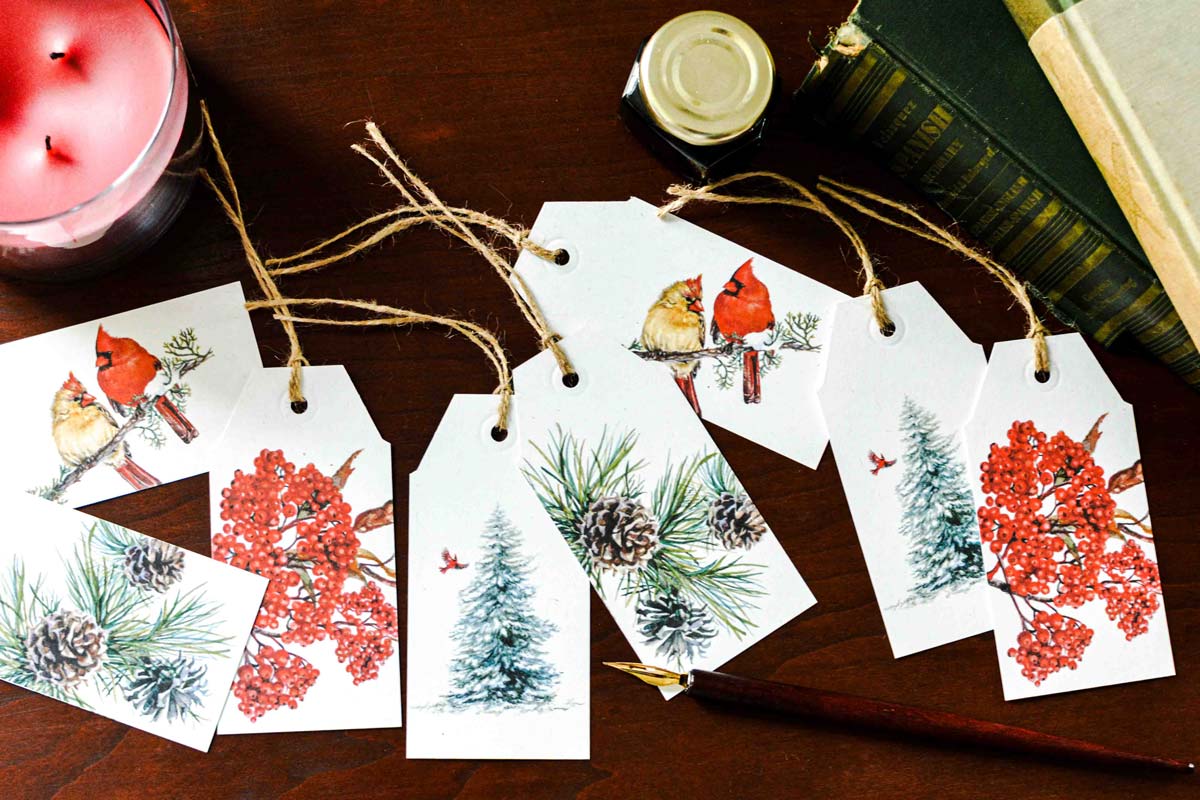 A set of white Christmas gift tags with rustic twine, printed with watercolor illustrations of cardinals, pine trees, pinecones, and red berries, arranged on a dark wooden tabletop with a pink candle, books, and a paintbrush nearby.