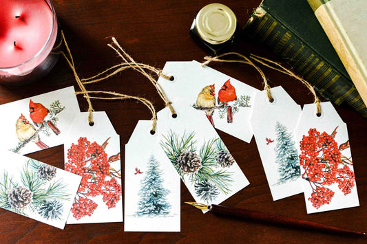 A set of white Christmas gift tags with rustic twine, printed with watercolor illustrations of cardinals, pine trees, pinecones, and red berries, arranged on a dark wooden tabletop with a pink candle, books, and a paintbrush nearby.