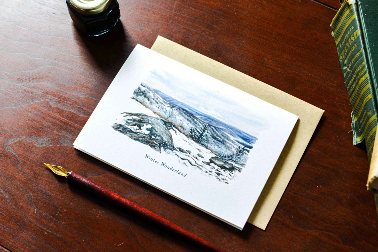 A watercolor greeting card titled 'Winter Wonderland' showing a snowy mountain landscape, displayed on a brown wooden desk next to a kraft envelope, fountain pen, and books.