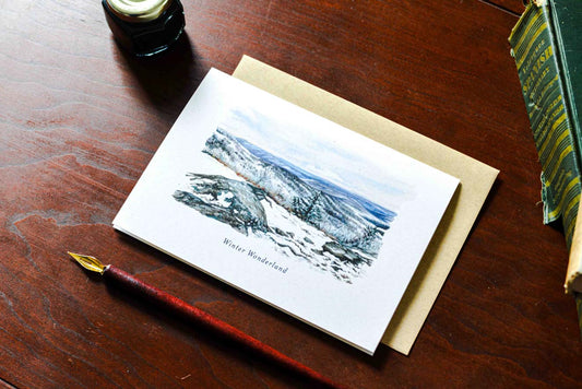 A watercolor greeting card titled 'Winter Wonderland' showing a snowy mountain landscape, displayed on a brown wooden desk next to a kraft envelope, fountain pen, and books.