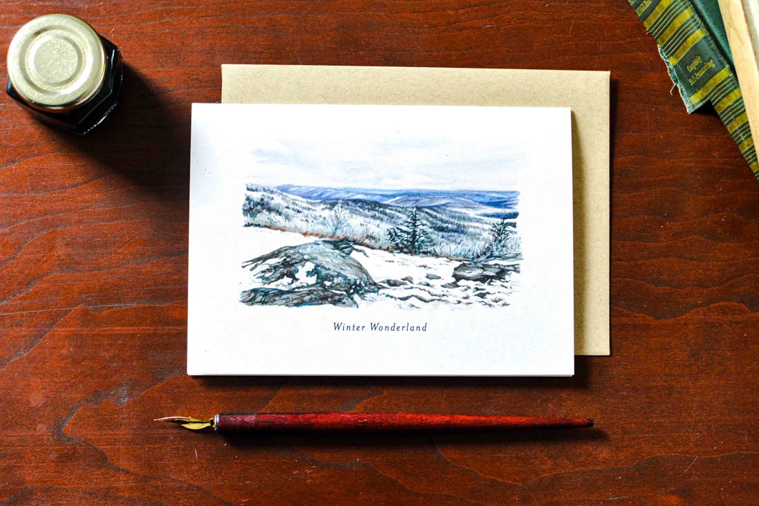 A greeting card with a watercolor landscape illustration labeled 'Winter Wonderland' lies on a brown wooden table, partially covered by a kraft envelope. A small ink bottle, a fountain pen nib, and a striped fabric edge are visible around it.