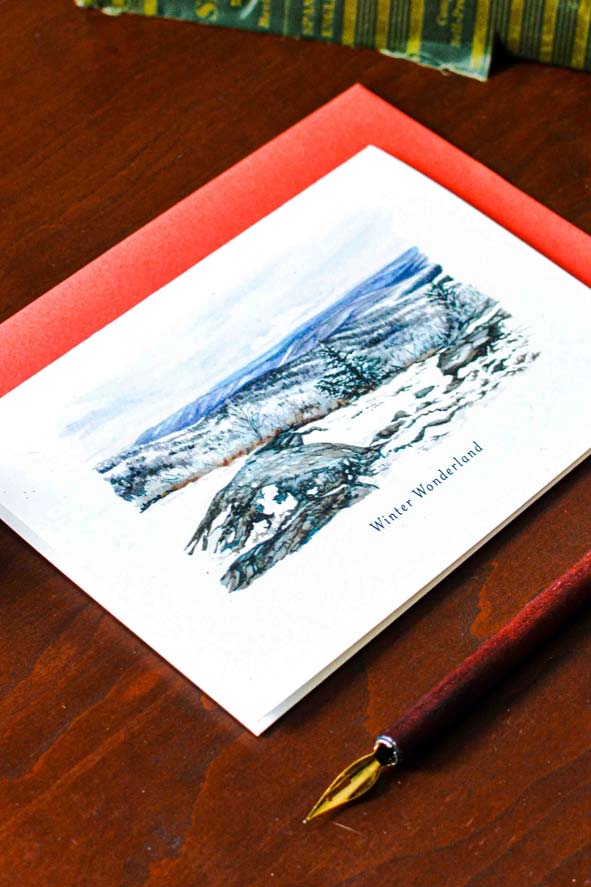 A watercolor greeting card titled 'Winter Wonderland' on a white cardstock, placed on a red envelope over a dark wooden table, with a fountain pen visible in the foreground.