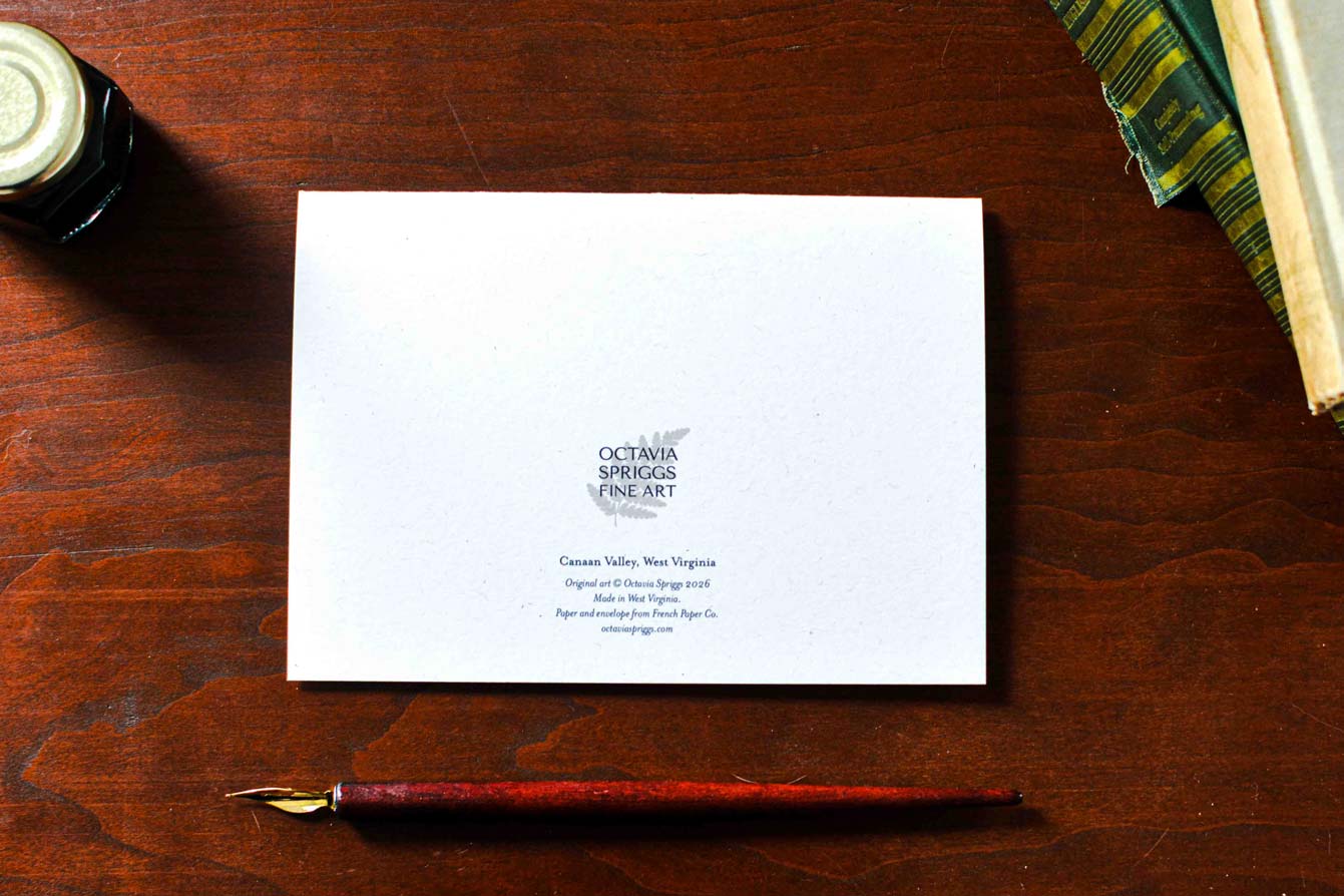 A white card backside with Octavia Springs Fine Art logo, displayed on a dark wooden desk alongside a fountain pen and books.